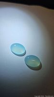Two oval, translucent Arctic Blue Chalcedony cabochon gemstones lying on a white surface under direct light, showcasing their smooth polished surfaces and soft blue glow.