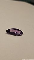 Close up image of a single deep purple marquis cut amethyst gemstone on a plain beige background showing light reflection and facets.
