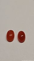 Pair of oval cabochon Mandarin Carnelian gemstones on a white background, showing their polished, smooth finish and orange-red color.