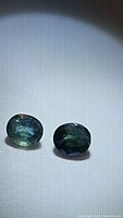 Two natural oval cut sapphires showing blue and green colors with visible inclusions, placed on a plain white background.