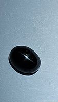 Close-up image of a polished oval cabochon jet black 4-star diopside gemstone showing the star-shaped reflection typical of this mineral under strong light.