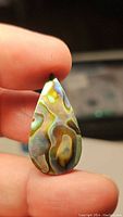 Close-up showing iridescent colors and natural swirling patterns on the teardrop abalone doublet gemstone held between fingers.