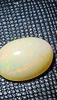 Close-up photo of an oval cabochon-cut Ethiopian opal showing a smooth polished surface with faint multicolored flashes on a textured black background.