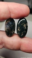 Close-up photo of two oval moss agate cabochons held between fingers showing natural moss-like inclusion patterns and polished surfaces.