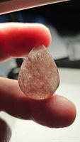 Close-up image of a pear-shaped cabochon Strawberry Quartz held between fingers showing pinkish tones and internal inclusions.