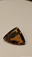Close-up image of a single deep brownish-orange Madeira citrine gemstone in a fancy trillion cut, resting on a plain surface.
