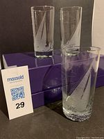 Three clear highball glasses each etched with a sailing motif, placed on purple boxes, visible etched sailboat and cloud designs.