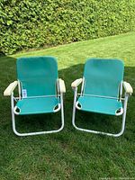 Two aqua beach chairs positioned outdoors on grass showing front view, white metal frames, and armrests.