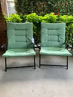 Two folding patio chairs side by side on a concrete porch in front of greenery, showing black frames and green cushions.