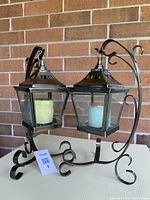 Photo showing two metal lanterns on decorative metal stands with candles inside, against a brick wall background.