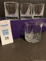 Four lowball glasses with etched sailing boat motifs, all placed on purple boxes with a lot card numbered 28 next to them.