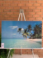 Large laminated print of a tropical beach scene featuring palm trees, clear blue water, and white sand, mounted on board and displayed on an easel against a brick wall.