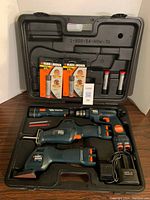 Full view of the open black plastic carrying case showing four Black & Decker cordless power tools (drill, flashlight, chop saw, sander), charger, and battery packs including two sealed batteries in packaging and two loose batteries.