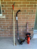 Photo of the three garden tools: pole pruner, craftsman weed wacker, and black & decker hedge trimmer against a brick wall.