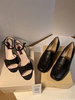 Two pairs of shoes on beige boxes, one black suede sandal with ankle strap and one black leather loafer with wedge sole, both size 37