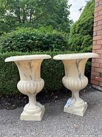 Two ivory marble-look resin garden urns standing on pavement outdoors, next to shrubbery and a brick wall.