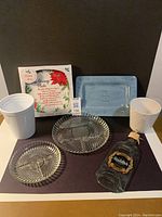 Photo showing all items in the lot: the Christmas Giving Plate in its box, two glass divided relish dishes, blue Live Laugh Love tray, two white flower pots, and Glenfiddich serving plate.