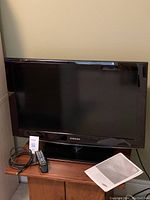 Samsung 32-inch LCD TV displayed on wooden stand with remote control and manual next to it and cable coiled beside remote.