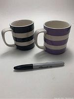 Front side view of two striped ceramic mugs, one black and one purple, with a Sharpie marker for scale.