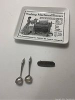 Photo showing two small pewter spoons, a wooden church pew number plaque marked '19', and a vintage metal ashtray with printed advertisement.