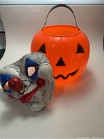 Orange plastic pumpkin blow mold candy bucket with carved face details and vintage rubber clown mask in front.