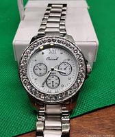 Men's silver-tone wristwatch with white dial, Roman numeral XII, and crystal-studded bezel shown on green textured background.