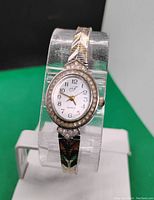 Front view of the women's quartz watch showing white oval face with Arabic numerals and decorative rhinestone bezel and metal band with gold and silver chevron pattern.