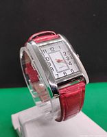 Front and slightly angled views of a women's rectangular silver-tone quartz watch with white face, black numerals, and red leather strap on a stand.