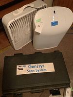 Three items together: Rabbit Air purifier, a white box fan, and black Genisys Scan System case on floor.