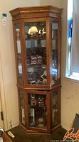 Full view of wooden corner display cabinet in a room corner showing glass doors and wood finish. Cabinet is filled with various items (not included).