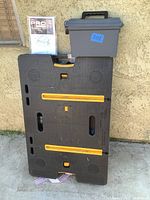 Folded black Keter work table leaning against stucco wall with yellow clamp rails; gray toolbox on top; instruction manual propped beside.
