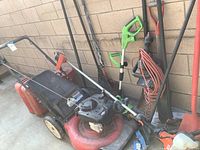 Overall photo showing the gas-powered lawn mower along with various gardening tools and gas tanks leaning against a brick wall.