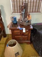 Dark wood side table with two drawers holding cowboy statue lamp, blue flashlight, and sunglasses. Lamp shade on carpet in front.