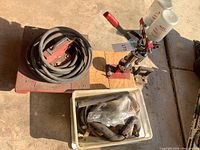 MEC lever-action pellet loader, dual primer feed bottles, coiled hose on metal plate, tub of plumbing fittings displayed on concrete surface