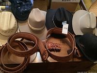 Multiple men's cowboy hats in black, cream felt, and natural straw colors along with brown leather belts and holsters on a wooden surface.