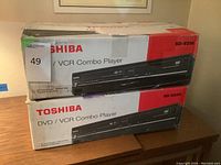 Two Toshiba DVD/VCR combo player boxes stacked on a wooden piece of furniture, clearly showing the Toshiba branding and model number SD-V296.