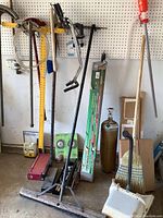 Photo of assorted tools including pipe wrench, lawn aerator, broom, floor jack, garden hose, gas cylinder, and other miscellaneous items hung on pegboard and set on floor.