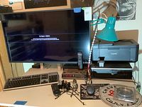 Photo showing Vizio 40-inch TV screen with remote on desk. HP keyboard and mouse in front, Canon TR4720 printer on the right, digital clock and blue-green desk lamp also on the desk.