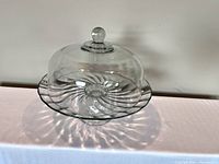 Side view of clear glass cake platter with swirl pattern and matching glass dome, displayed on a flat surface against white background.
