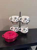 Four white ceramic mugs with black snowflakes arranged in a black wrought iron holder beside a pink stoneware mini brie baker with lid on a dark surface.