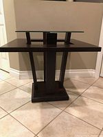 Front view of the modern end table showing the wooden base and glass top with the table standing on a tiled floor against a wall.