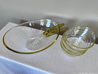 Clear acrylic large oval salad bowl with yellow rim, two serving utensils resting inside, and four smaller matching bowls stacked beside it on a white surface against a plain background.