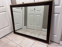Front view of the large rectangular mirror leaning against white paneled doors, showing dark brown frame and clear reflective glass surface.