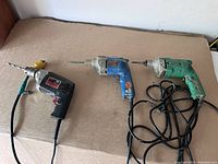 Overview of three electric drills: Hitachi (green), Ryobi (blue), Skil (black) positioned on surface showing cords and drill bits.