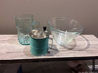 Photo shows glass water pitcher, vintage metal flour sifter with crank handle, and one large clear glass mixing bowl arranged on wooden surface.