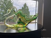 Full side profile of vintage glass swan figurine showcasing clear glass neck and beak with flowing green and brown swirled body placed on a curved black wooden surface in front of window with natural light.