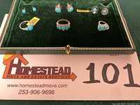 Green velvet jewelry box with three turquoise sterling silver rings and three pairs of turquoise pierced earrings on display, including tags on some items.