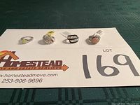 Four sterling silver rings displayed on a card with seller and lot number details, showing each ring's front view and stone colors.