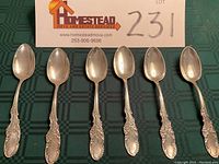 Photo shows the full lot of six ornate sterling silver teaspoons laid out horizontally, showing front side with bowls and handle design