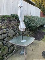 Full view of the glass top patio table with closed beige folding umbrella and umbrella stand on outdoor concrete surface with stone wall background.